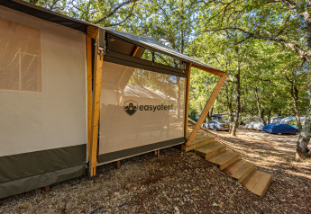 Tienda safari con escalera de madera en un camping, rodeada de árboles y coches bajo luz filtrada.