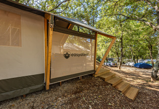 Safari tent with wooden stairs at a campground, surrounded by trees and vehicles, in filtered sunlight.