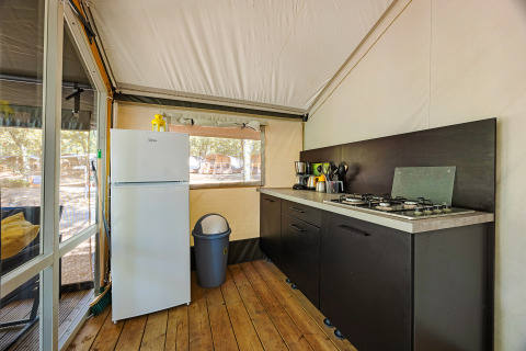 Interior view of a safari tent kitchen with stove, refrigerator, trash bin and wooden flooring.