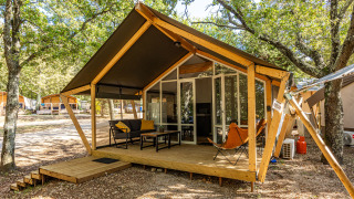 Tienda safari con terraza de madera y asientos al aire libre en un campamento sombreado por árboles.