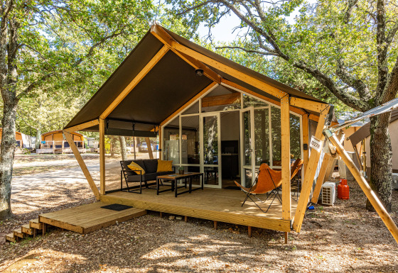 Tienda safari con terraza de madera y asientos al aire libre en un campamento sombreado por árboles.