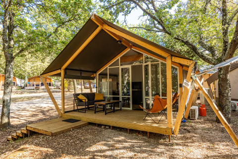 Tienda safari con terraza de madera y asientos al aire libre en un campamento sombreado por árboles.