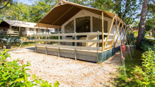 Tienda safari con porche, instalaciones sanitarias y aire acondicionado, rodeada de árboles verdes.