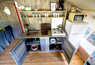 Pequeña cocina en una tienda safari con estanterías abiertas, fogón, nevera, microondas y utensilios.
