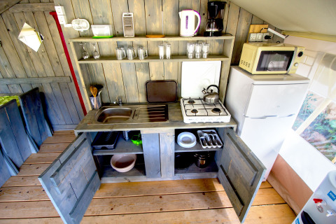 Pequeña cocina en una tienda safari con estanterías abiertas, fogón, nevera, microondas y utensilios.