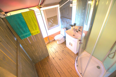 Bagno in tenda safari con pavimento in legno, doccia, wc, lavabo e asciugamani colorati a Camping Valkanela.