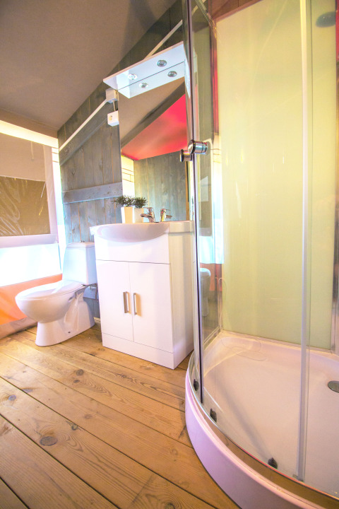Baño en una tienda safari con suelo de madera, cabina de ducha, inodoro y mueble blanco de lavabo.
