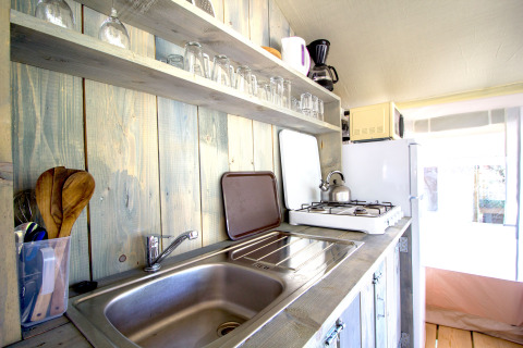 Kitchen area in a safari tent with a sink, stove, wooden utensils, glasses, and a fridge in sunlight.