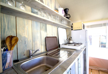 Kitchen area in a safari tent with a sink, stove, wooden utensils, glasses, and a fridge in sunlight.
