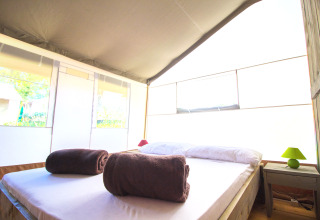 Interior luminoso de tienda safari con cama doble, dos toallas marrones enrolladas y mesa de noche con lámpara.