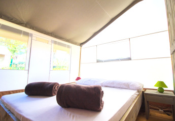 Camera da letto di tenda safari luminosa con letto matrimoniale, due asciugamani marroni e comodino con lampada.