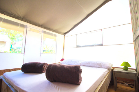 Interior luminoso de tienda safari con cama doble, dos toallas marrones enrolladas y mesa de noche con lámpara.