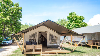Tenda safari moderna con veranda coperta, sedie da esterno e alberi verdi tutto intorno sotto il cielo blu.