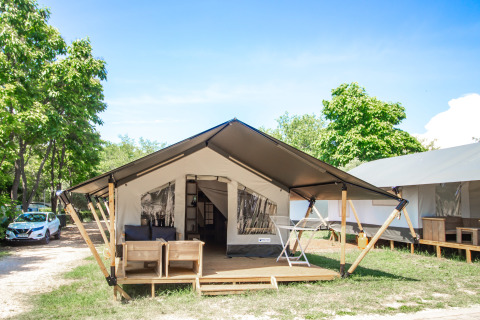 Tenda safari moderna con veranda coperta, sedie da esterno e alberi verdi tutto intorno sotto il cielo blu.