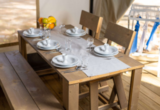 Mesa de comedor puesta en una tienda safari, con bancos de madera, platos blancos y bandeja de frutas.