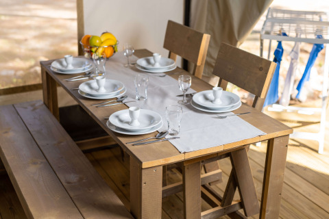Mesa de comedor puesta en una tienda safari, con bancos de madera, platos blancos y bandeja de frutas.