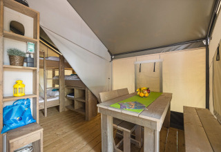 Interno di una tenda safari con letti a castello, tavolo da pranzo in legno, panche e scaffali.
