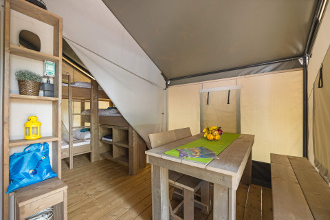 Interno di una tenda safari con letti a castello, tavolo da pranzo in legno, panche e scaffali.