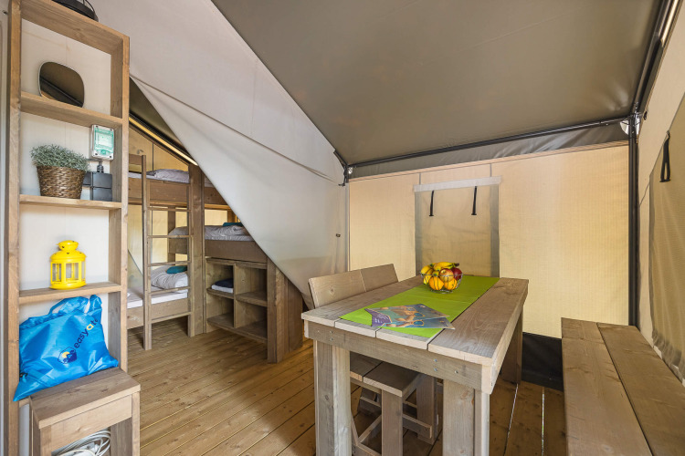 Interieur van een safaritent met stapelbedden, houten eettafel met banken en een open kast.