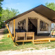 Safari Tent Comfort med en træveranda, udendørs stole og grønne omgivelser under klare himmel