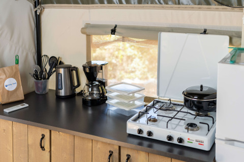 Køkkenbord i safaritelt med elkedel, kaffemaskine, gasblus, gryder, køkkenredskaber og plastikbokse.