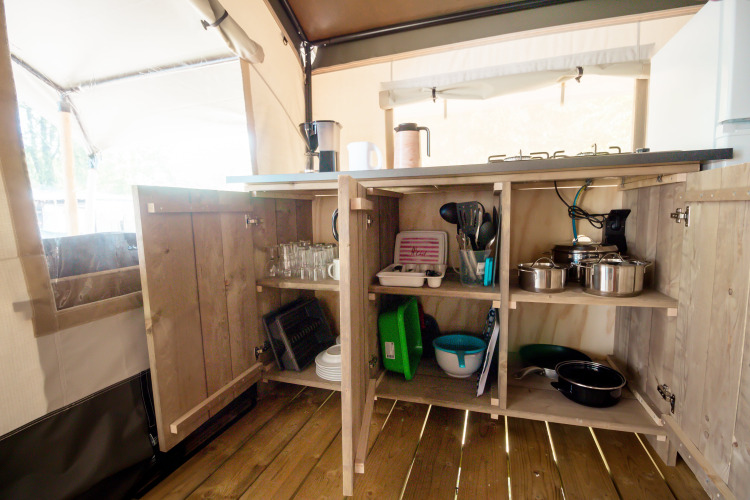 Placards de cuisine ouverts dans une tente safari, avec casseroles, vaisselle et verres sur un plancher en bois.