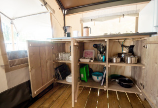 Open kitchen cabinets in a safari tent, stocked with pots, pans, dishes, and glasses on wooden flooring.