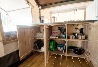 Open kitchen cabinets in a safari tent, stocked with pots, pans, dishes, and glasses on wooden flooring.