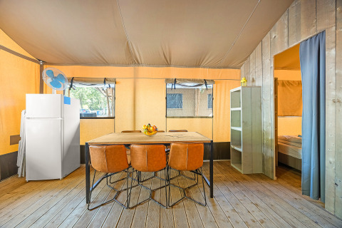 Innenraum eines Safari-Zeltes mit Holzboden, Esstisch, vier Stühlen, Kühlschrank und Schlafzimmerzugang.