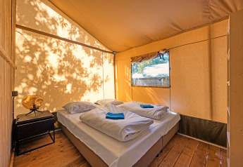 Interieur van een safaritent met tweepersoonsbed, nachtkastje en natuurlijk licht door het raam.
