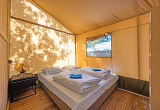 Vista interior de una tienda safari con cama doble, mesa lateral y luz natural entrando por la ventana.