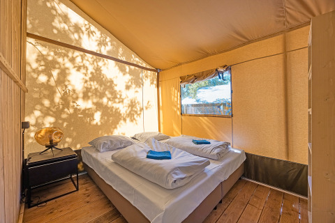 Interieur van een safaritent met tweepersoonsbed, nachtkastje en natuurlijk licht door het raam.