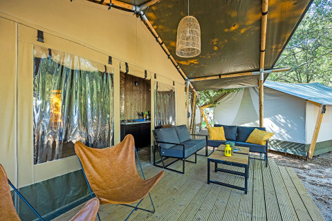 Terrasse de tente safari avec sièges confortables, fauteuils papillon et lanterne, dans un cadre boisé.
