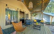 Safari tent veranda met comfortabele zitplaatsen, vlinderstoel en lantaarn, omgeven door bosrijke natuur.