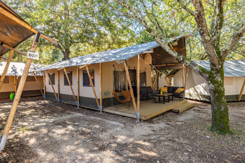 Vue extérieure d'une tente Safari Lodge climatisée avec terrasse en bois et coin salon au milieu des arbres.