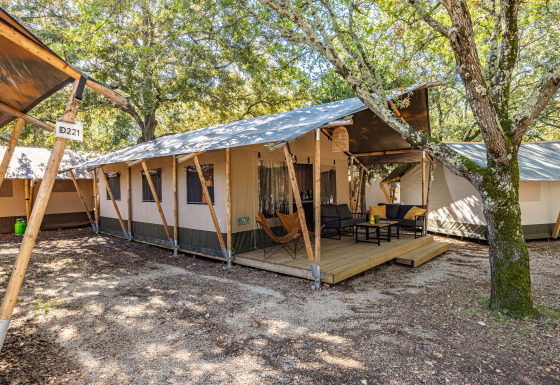 Vue extérieure d'une tente Safari Lodge climatisée avec terrasse en bois et coin salon au milieu des arbres.