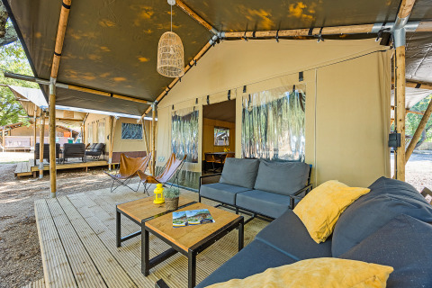 Zona de estar exterior de tienda safari con sofás, mesas y vista a otras tiendas en el campamento.