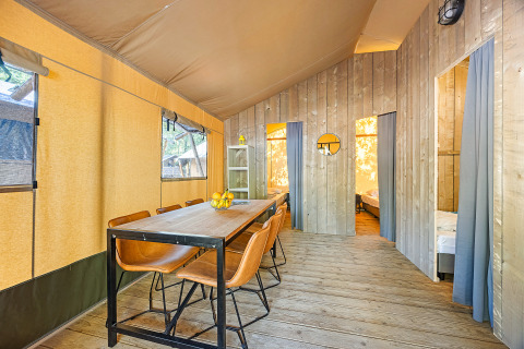 Vista interior de una tienda safari con mesa de comedor, sillas de cuero y dos dormitorios divididos por cortinas.