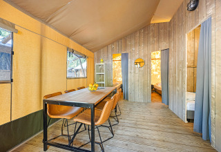 Vista interior de una tienda safari con mesa de comedor, sillas de cuero y dos dormitorios divididos por cortinas.