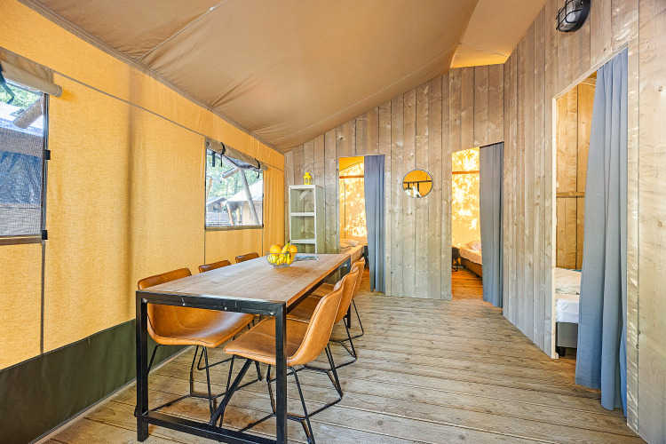 Interior view of a safari tent with dining table, leather chairs, and two bedrooms separated by curtains.