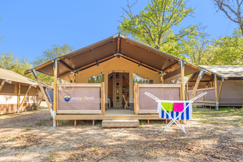 Safari-Zelt Lodge mit Klimaanlage, umgeben von Bäumen, bunte Handtücher hängen vorne am Geländer.