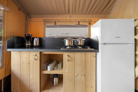 Kleine Küchenzeile im Safarizelt mit Kühlschrank, Herdplatten, Kaffeemaschine und Holzmöbeln.