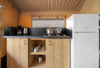 Pequeña cocina dentro de una tienda safari con nevera, fogones, cafetera y armarios de madera con vasos.