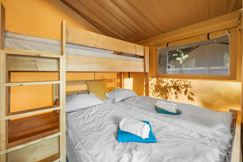 Vista interior de una tienda safari con cama doble, litera de madera, ventana grande y toallas dobladas.