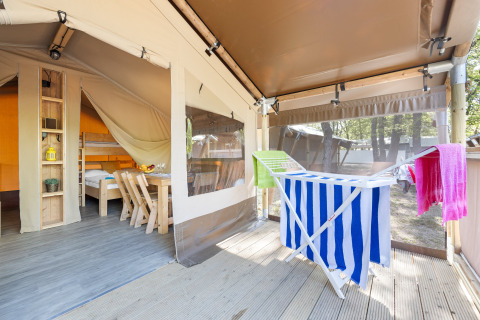 Bright safari tent interior with wooden furniture, drying rack holding colorful towels, forest outside.