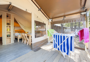 Bright safari tent interior with wooden furniture, drying rack holding colorful towels, forest outside.
