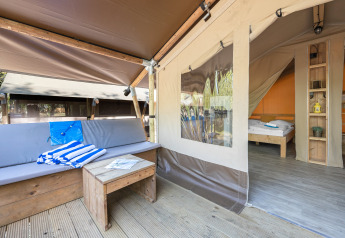 Safari tent featuring a cozy outdoor lounge with wooden furniture and view into the bedroom with a bed.