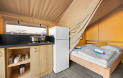 Interior of a safari tent featuring a kitchenette, fridge, and double bed with neatly folded towels.