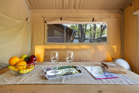 Inside a safari tent with a fruit basket, two wine glasses on a tray, papers on a rustic wooden table.