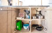 Küchenschrank im Safari Tent Comfort mit Töpfen, Utensilien, Geschirr und Besteck auf Regalen.